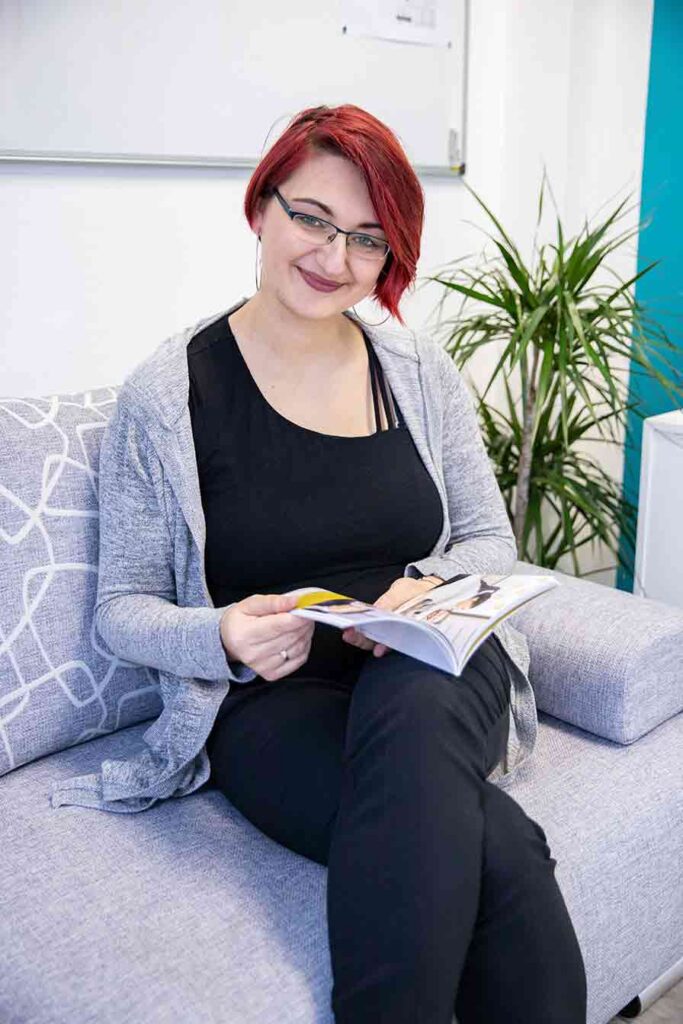 Alin_gruender_business Alin Gruender sitzt auf einem grauen Sofa, mit Buch in der Hand. Sie lächelt freundlich in die Kamera.